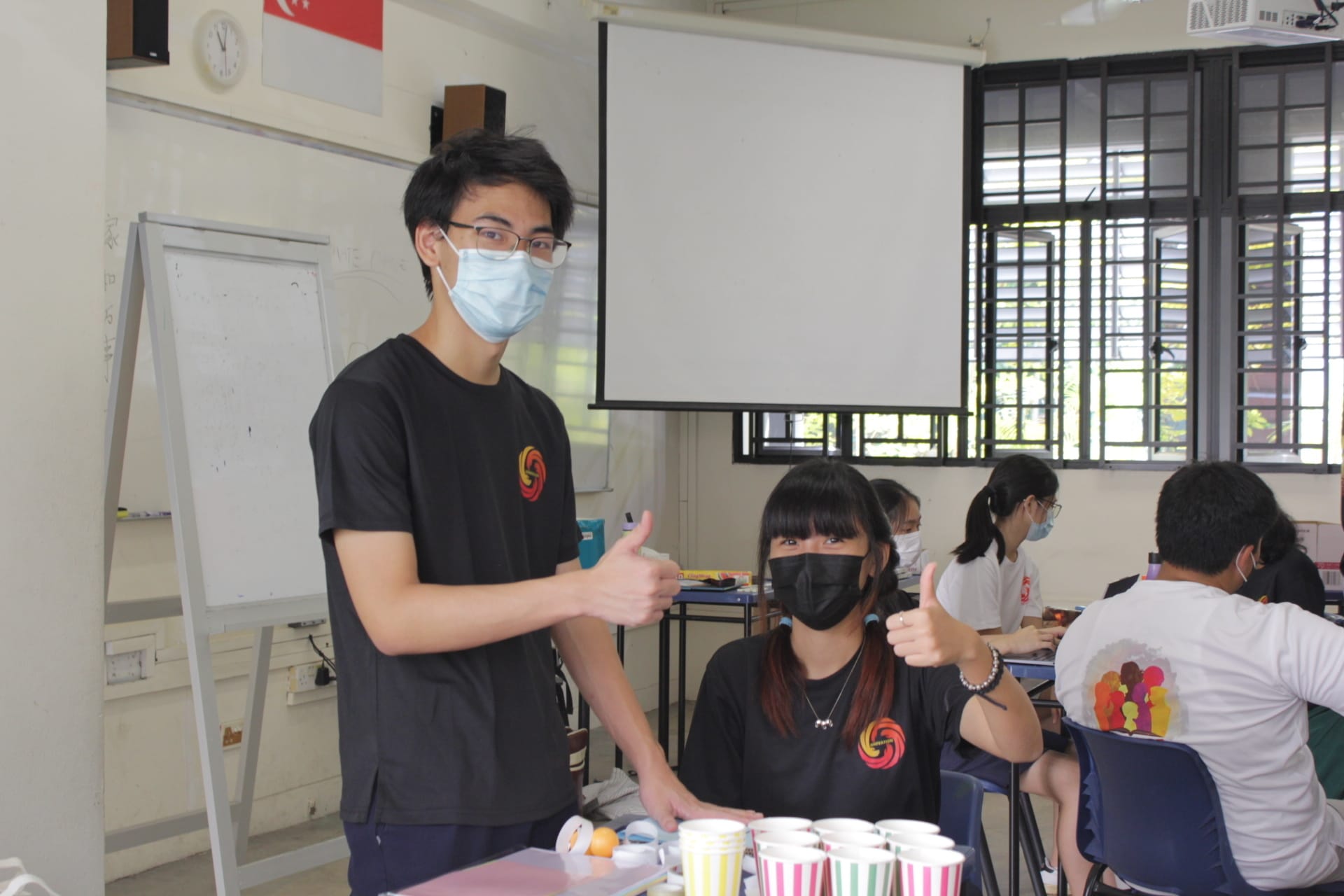 Two masked students in black shirts give thumbs up in a classroom with a projector screen and other students seated nearby.
